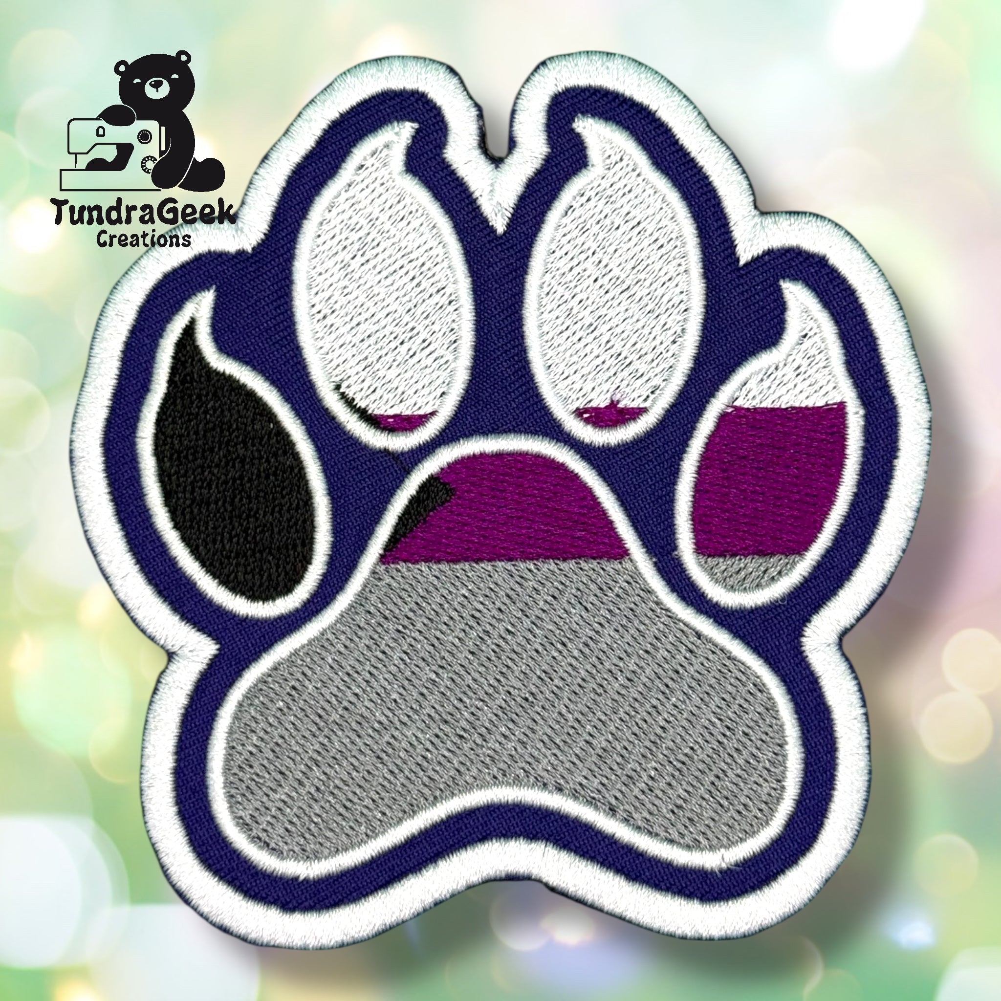 An embroidered paw-shaped patch in demisexual pride flag colors — black, gray, white, and purple/magenta stripes. The patch has a deep navy background with white border edging and inner pad detail. One toe bean is solid black.