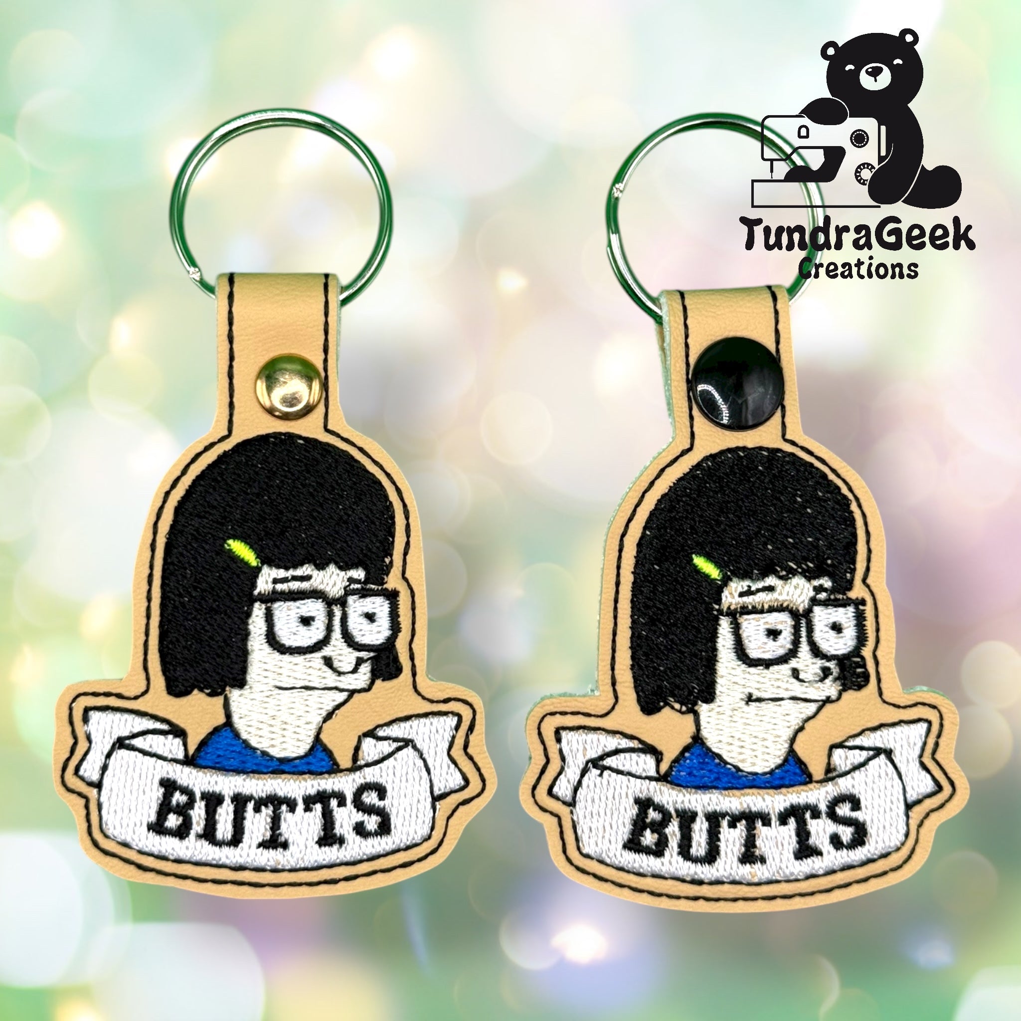 Two embroidered keychains featuring a cartoon character with glasses and black hair, labeled “BUTTS” below, with the TundraGeek Creations logo in the top right corner.