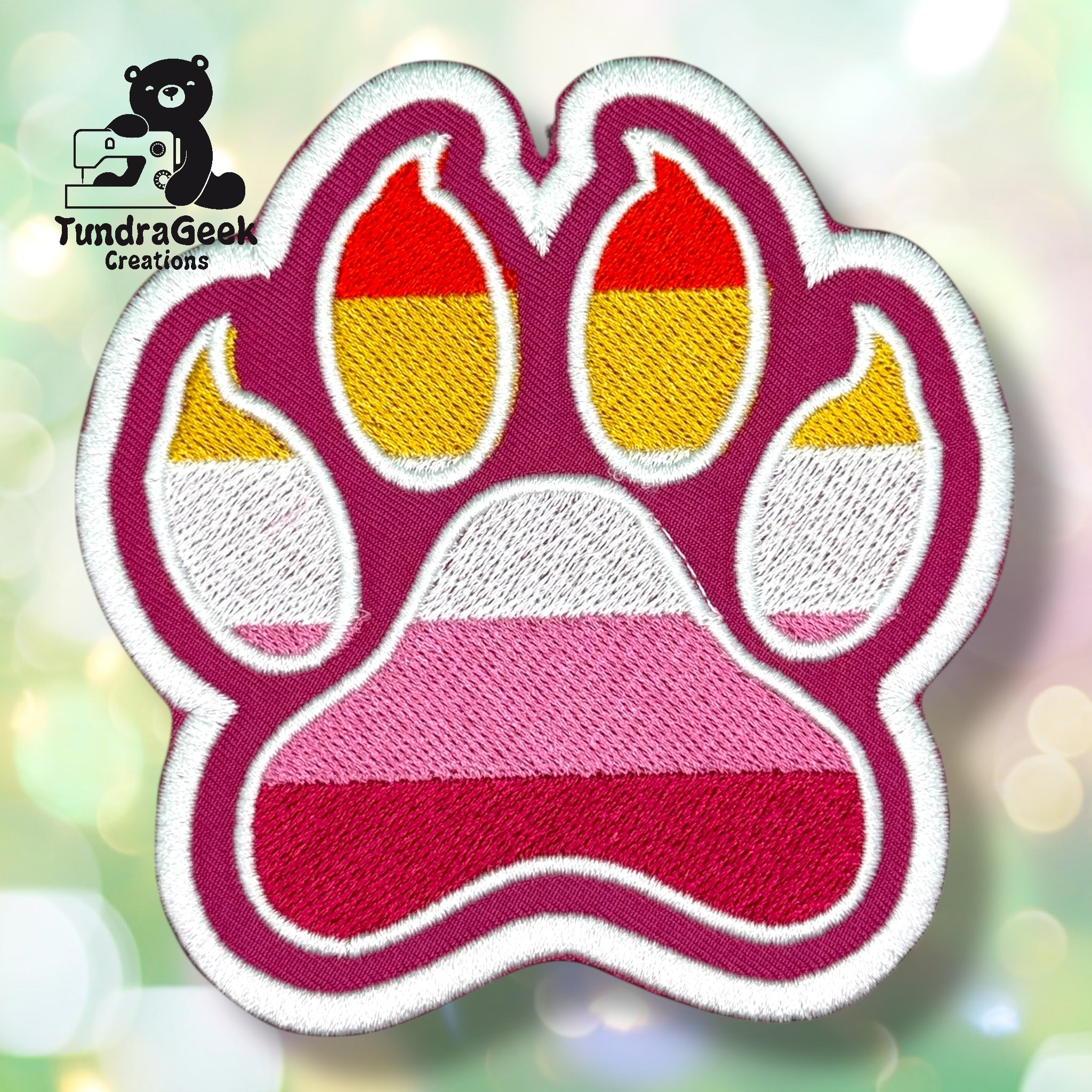 An embroidered paw-shaped patch in lesbian pride flag colors — horizontal stripes of dark magenta/red, orange-red, gold/yellow, white, pink, and deep pink/red. The patch has a magenta background with white border edging and inner pad detail.