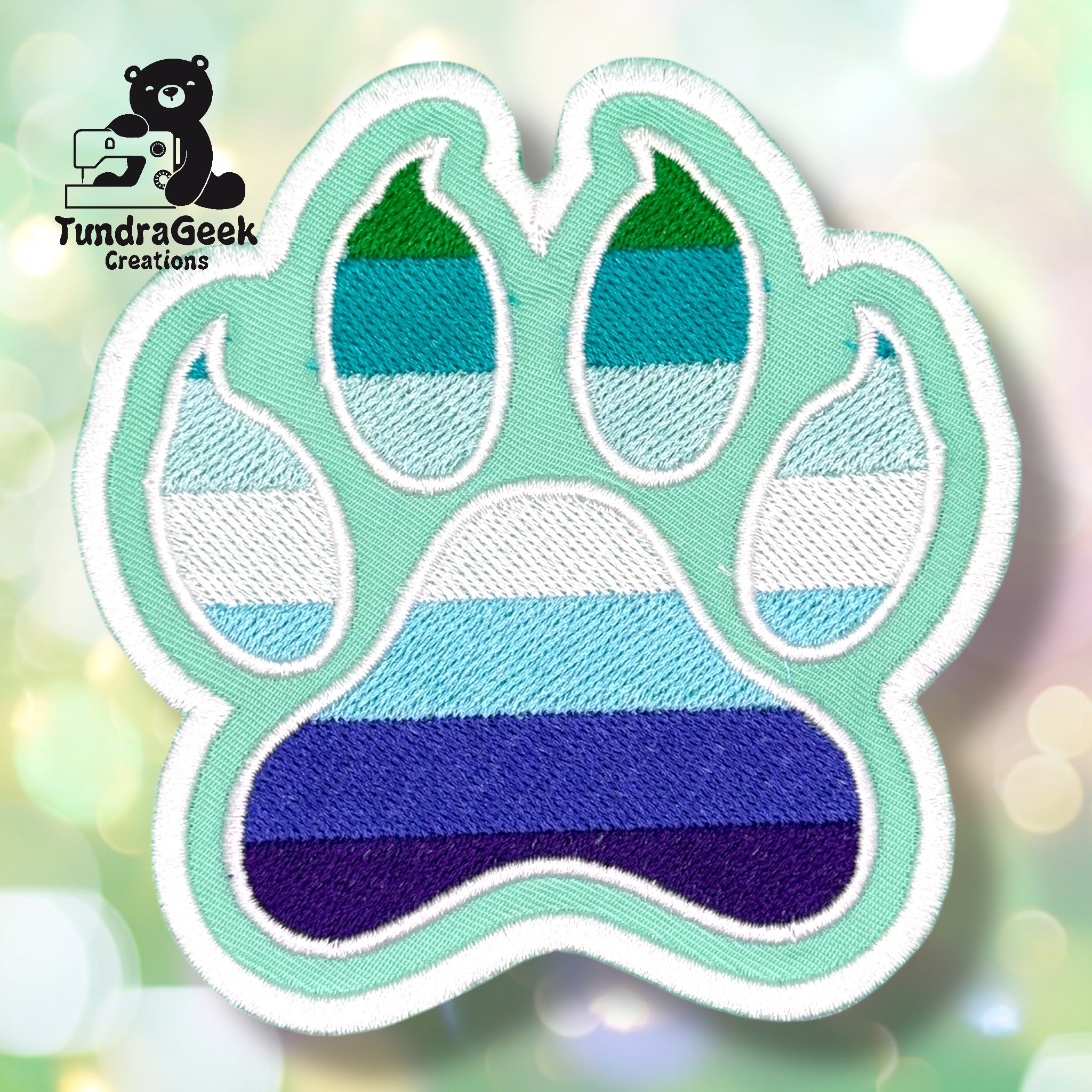 An embroidered paw-shaped patch in MLM pride flag colors — horizontal stripes of dark green, teal, light teal, white, light blue, periwinkle/blue, and dark indigo. The patch has a mint/seafoam green background with white border edging