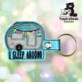 Blue camper keychain with I SLEEP AROUND text, colorful flag bunting, and heart details. TundraGeek Creations logo with a bear holding a sewing machine in the top right corner.