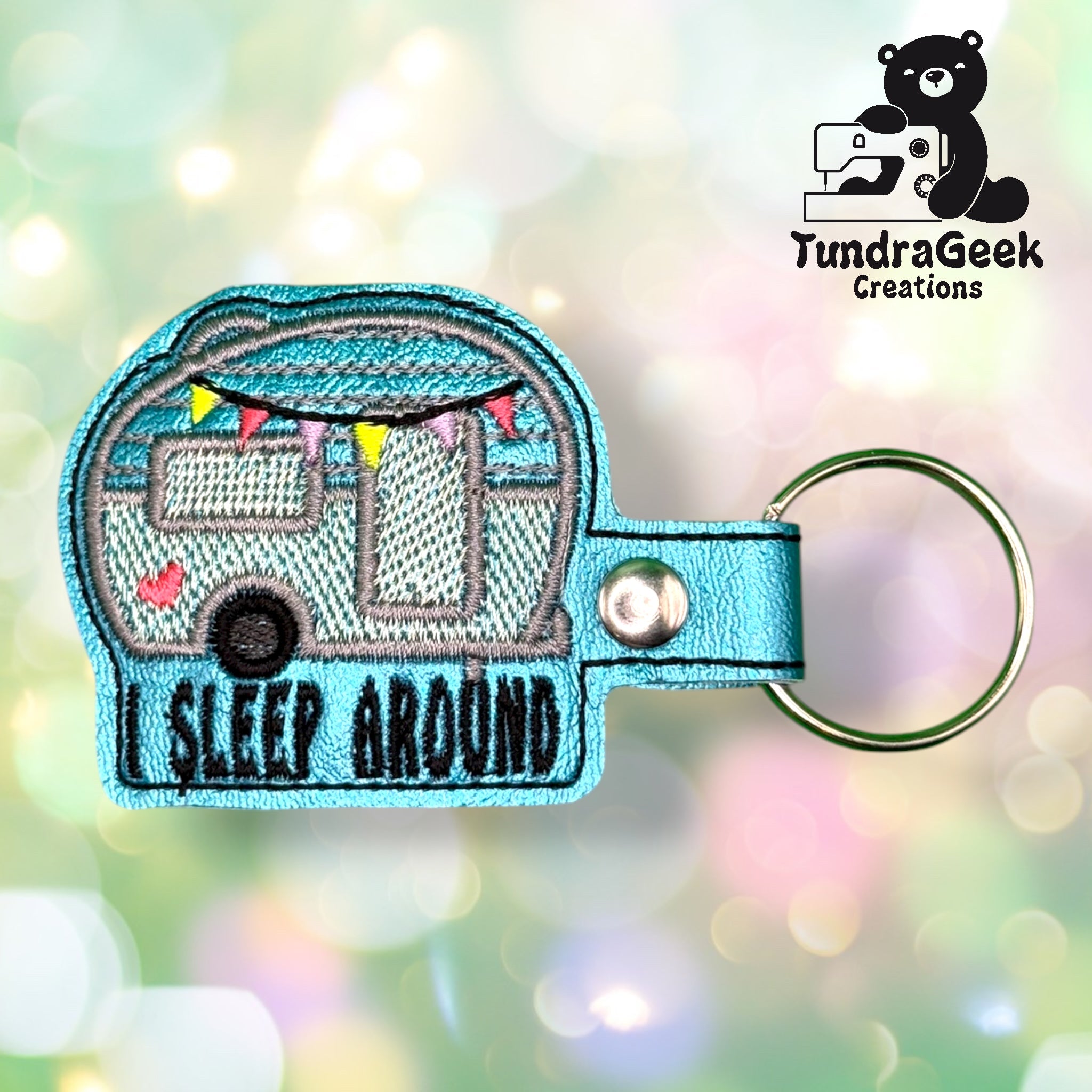 Blue camper keychain with I SLEEP AROUND text, colorful flag bunting, and heart details. TundraGeek Creations logo with a bear holding a sewing machine in the top right corner.