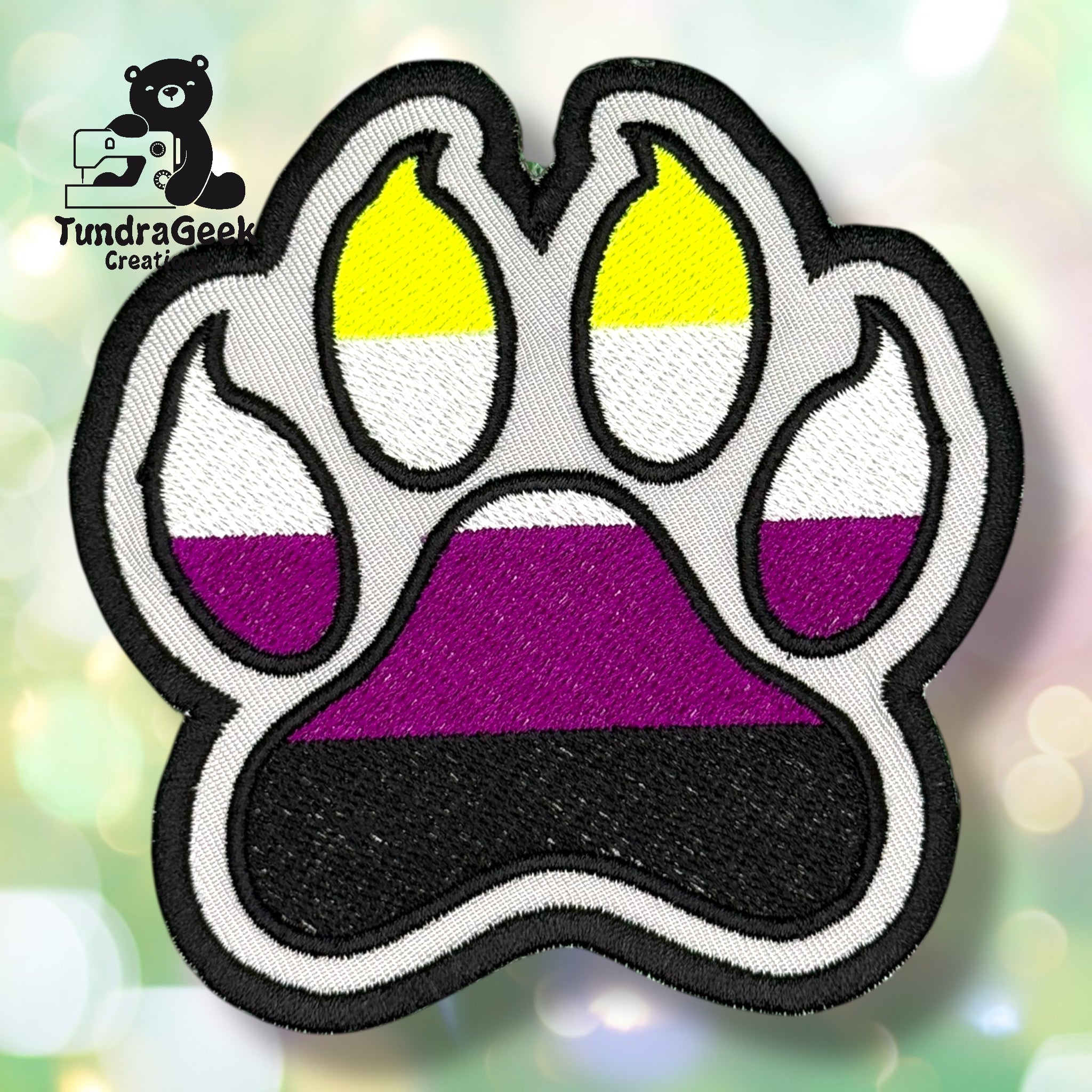 An embroidered paw-shaped patch in non-binary pride flag colors — horizontal stripes of yellow, white, purple, and black. The patch has a black background with white border edging. Toe bean tips are bright yellow