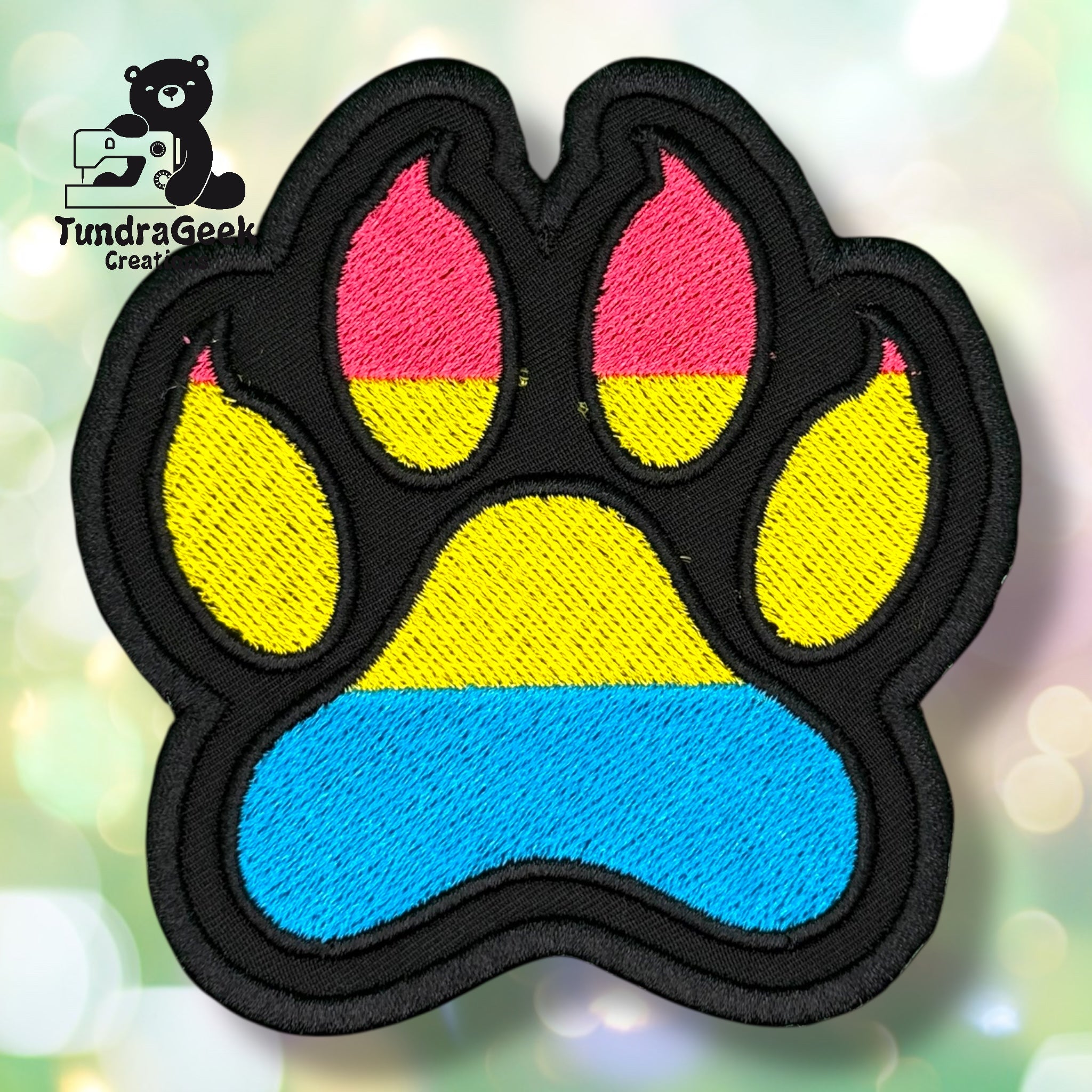 An embroidered paw-shaped patch in pansexual pride flag colors — horizontal stripes of hot pink, yellow, and sky blue. The patch has a solid black background and border, with the flag colors filling the paw pad and toe bean areas
