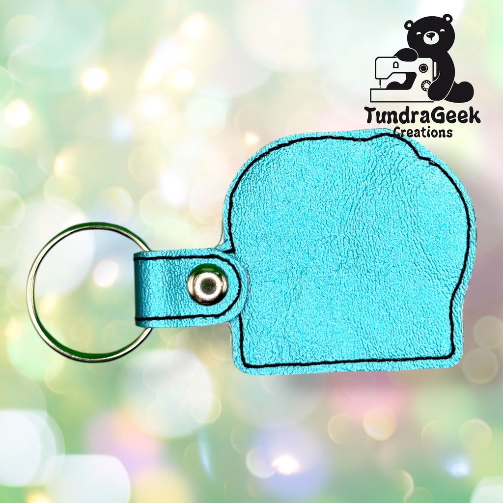 Blue faux leather keychain shaped like an elephants head with a metal ring, against a blurred colorful background. Logo reading TundraGeek Creations with a bear and sewing machine in the top right.