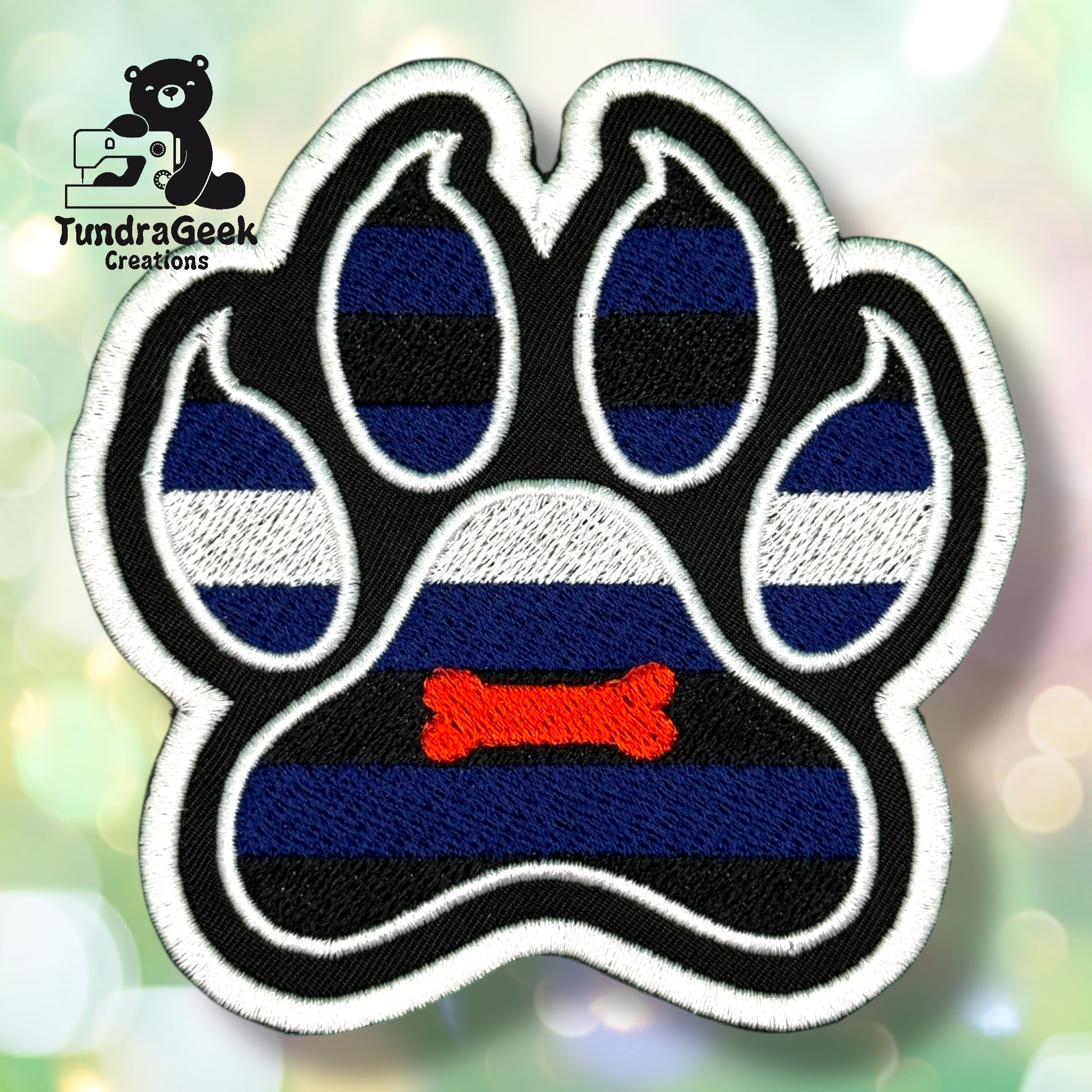 An embroidered paw-shaped patch in pup pride flag colors — alternating horizontal stripes of royal blue and black with a white stripe across the middle. A small orange embroidered bone sits in the center pad area. White border edging on a black background.