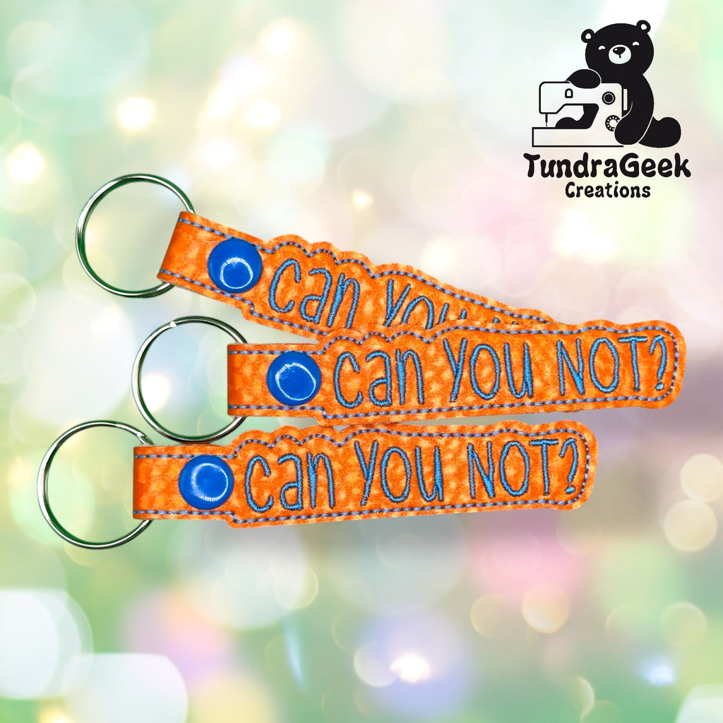Three orange keychains with blue stitching each read can you not? The background is blurred with light spots. TundraGeek Creations logo with a bear and sewing machine is in the upper right corner.