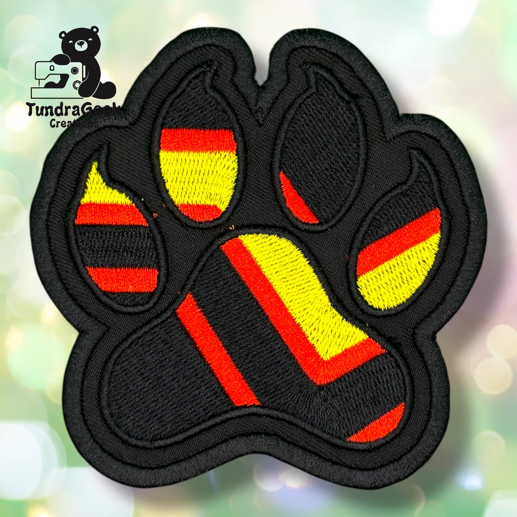 An embroidered paw-shaped patch in rubber/latex pride flag colors — bold diagonal or chevron stripes of black, red, and yellow on a solid black background and border. The high-contrast design gives the patch a striking, graphic appearance.