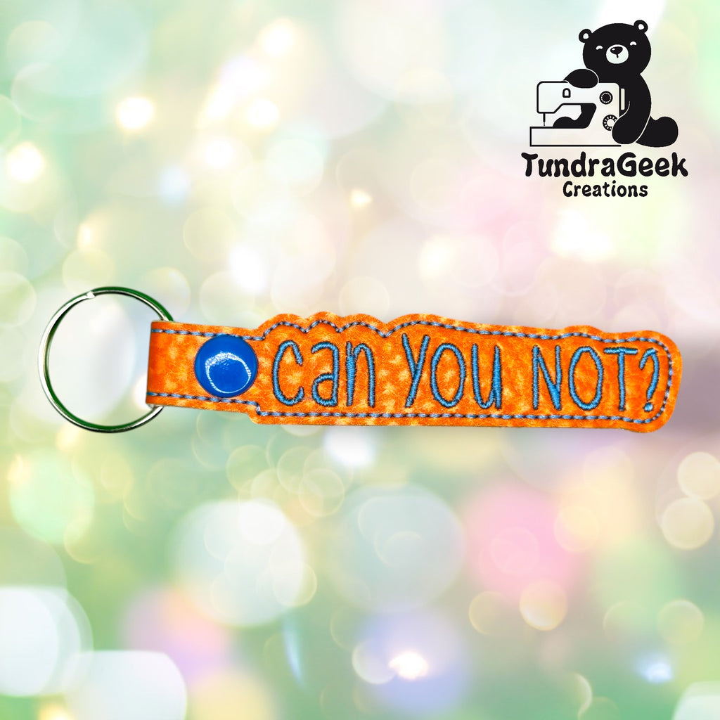 An orange embroidered keychain with the text CAN YOU NOT? in blue letters, displayed on a blurred pastel background with the TundraGeek Creations logo in the top right corner.