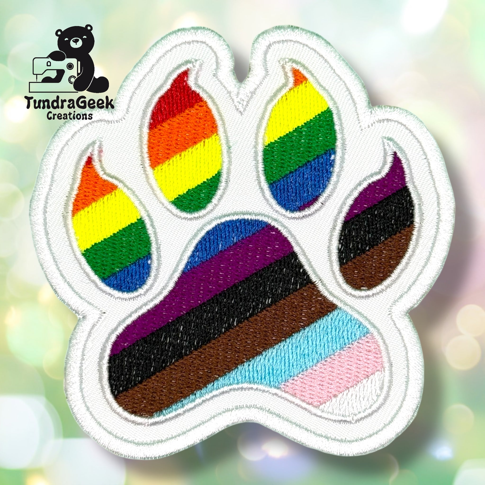 An embroidered paw-shaped patch featuring the Progress Pride flag design — a white background with a diagonal chevron of light blue, pink, white (trans flag colors), brown, and black, overlaid on classic rainbow stripes of red, orange, yellow, green, blue, and purple. White border edging.