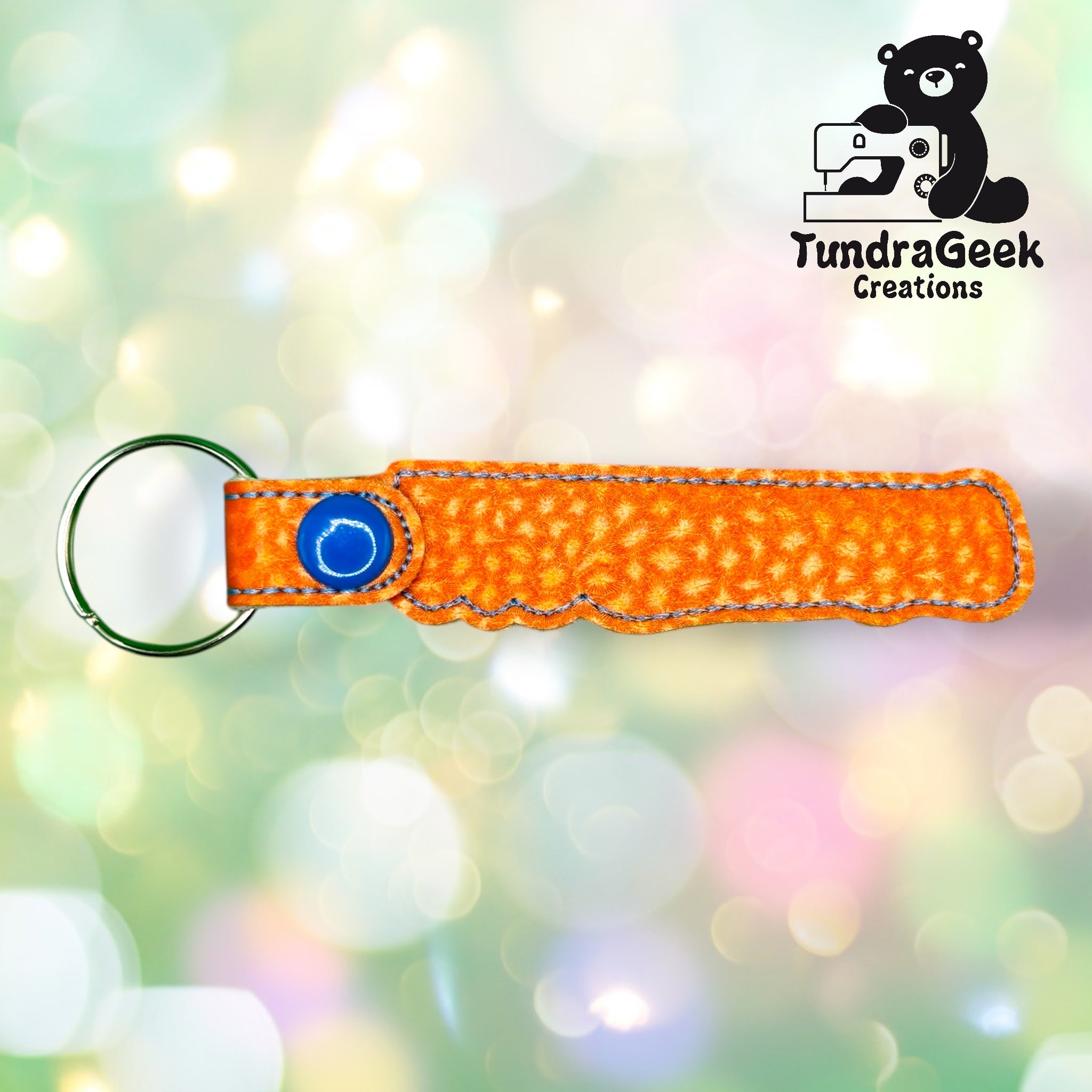 An orange textured fabric keychain with a blue snap button and a metal ring, displayed on a blurred pastel background with the TundraGeek Creations logo in the top right corner.