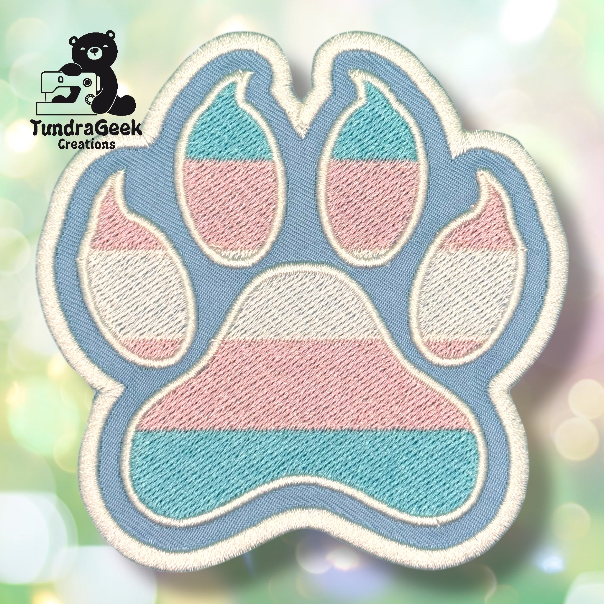 An embroidered paw-shaped patch in transgender pride flag colors — horizontal stripes of light teal/blue, pink, and white. The patch has a soft periwinkle blue background with cream/white border edging and inner pad detail.
