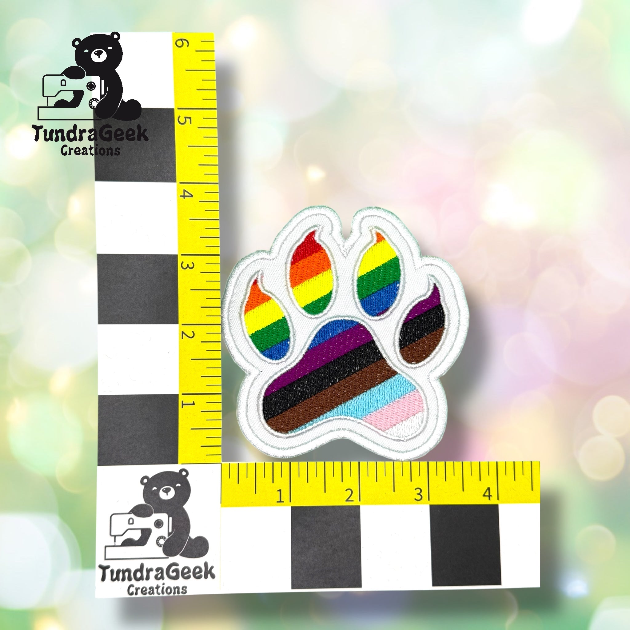 An embroidered paw-shaped patch featuring the Progress Pride flag design — a white background with a diagonal chevron of light blue, pink, white (trans flag colors), brown, and black, overlaid on classic rainbow stripes of red, orange, yellow, green, blue, and purple. White border edging. Sitting next to a yellow, black, and white scale ruler.