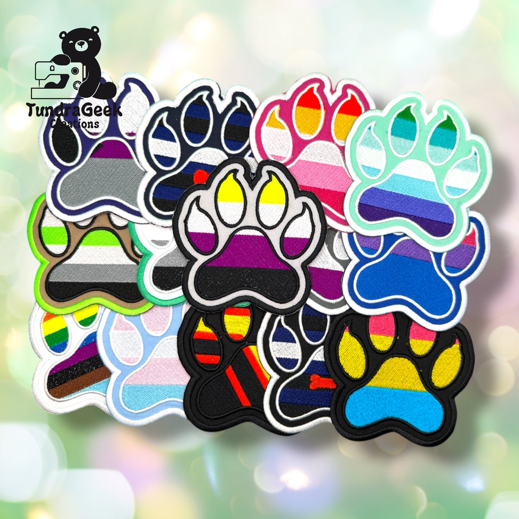 A collection of approximately fifteen embroidered paw-shaped pride patches spread out on a soft bokeh background. The patches display a wide variety of pride flag color combinations including rainbow, bisexual, pansexual, non-binary, asexual, leather, agender, progress pride, and others. Each patch features bold embroidered flag stripes within a paw print shape with a contrasting outline.
