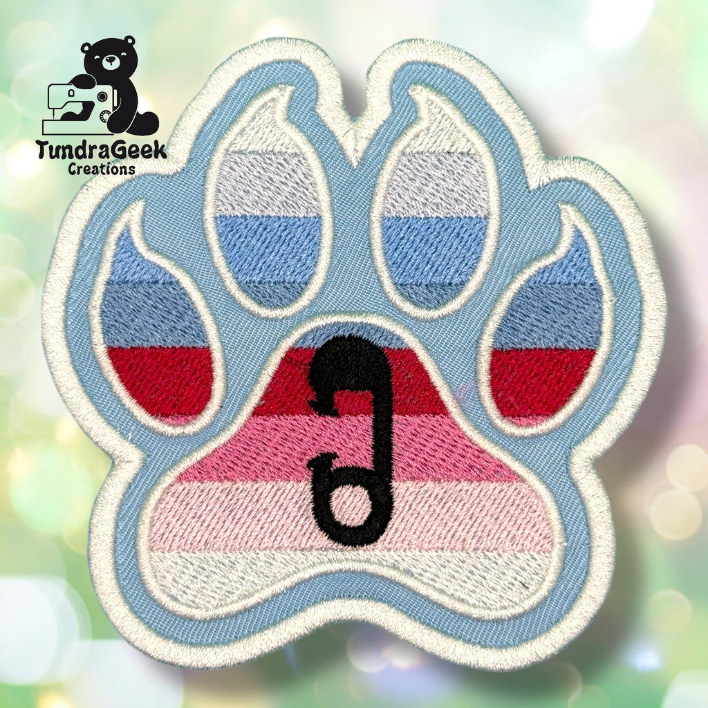 An embroidered paw-shaped patch in the colors of the ABDL pride flag — horizontal stripes of light blue, periwinkle, white, red, and pink. A small black safety pin is embroidered in the center pad area. The patch has a cream-colored outline on a light blue background.