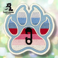 An embroidered paw-shaped patch in the colors of the ABDL pride flag — horizontal stripes of light blue, periwinkle, white, red, and pink. A small black safety pin is embroidered in the center pad area. The patch has a cream-colored outline on a light blue background.