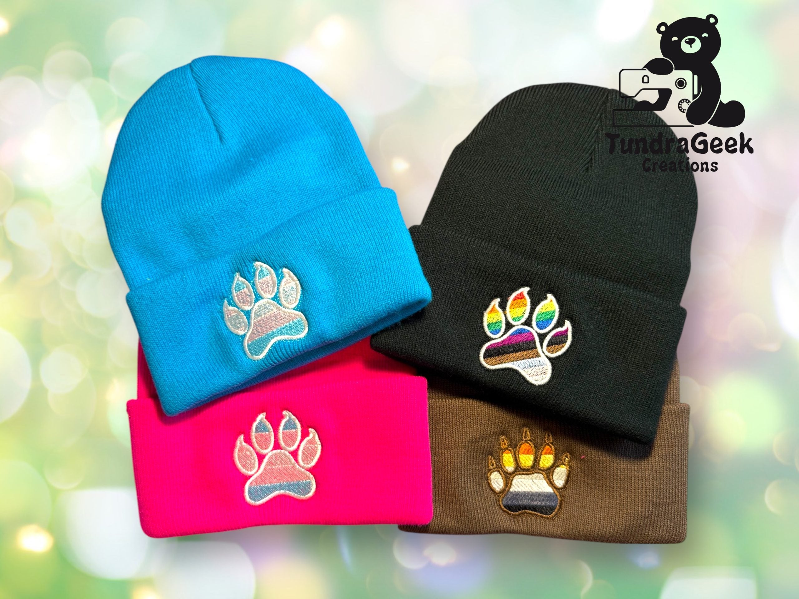 Four beanies in blue, black, pink, and brown, each featuring an embroidered paw print design. TundraGeek Creations logo is in the top right corner.