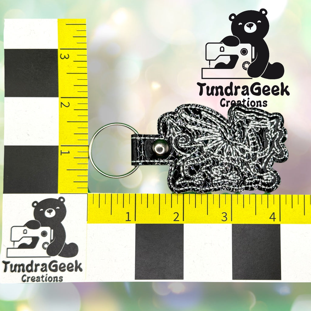 A black and white dragon keychain is shown next to a ruler measuring approximately 2 inches wide, with the TundraGeek Creations logo in the background.
