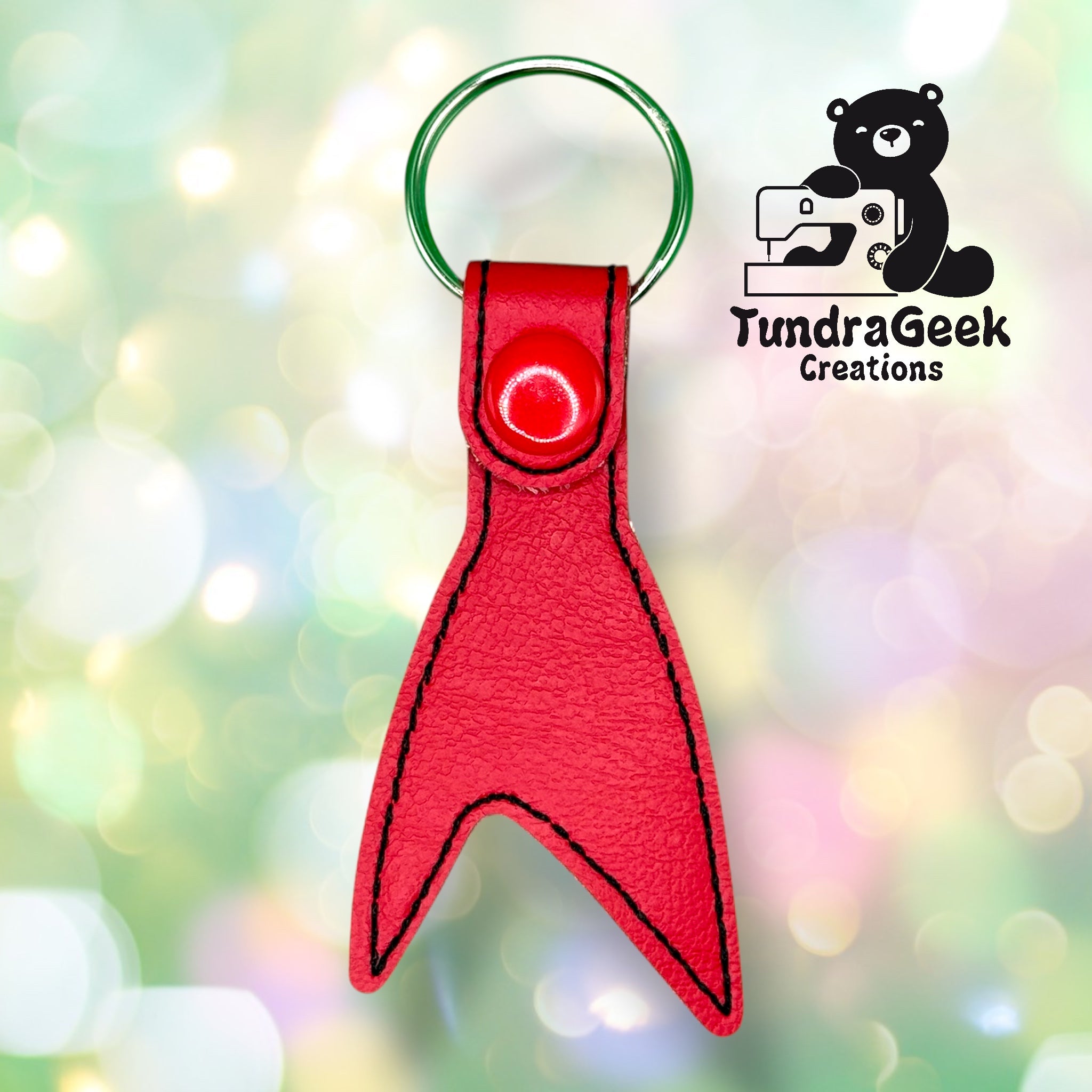 A red, leather keychain in the shape of a fish tail with black stitching, a green metal keyring, and the TundraGeek Creations logo on a blurred pastel background.