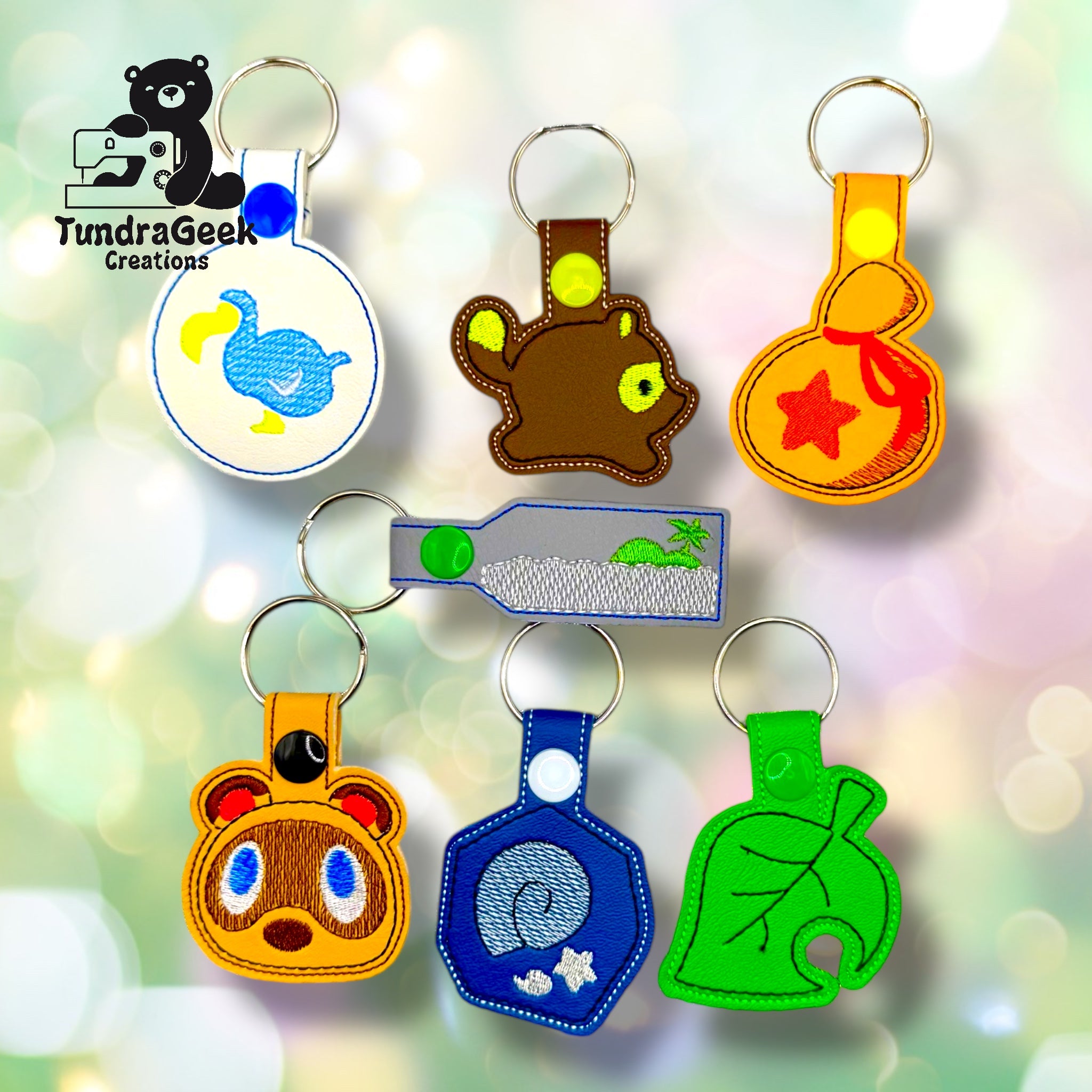 A collection of seven colorful embroidered keychains with various animal and object designs, displayed on a soft pastel background with a TundraGeek Creations logo in the corner.
