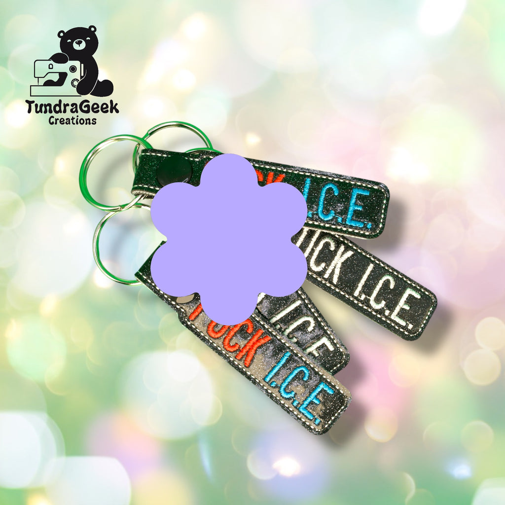Three rectangular keychains with the words F*CK ICE in colorful letters are displayed on a blurred pastel background. A purple flower shape partially obscures the text.