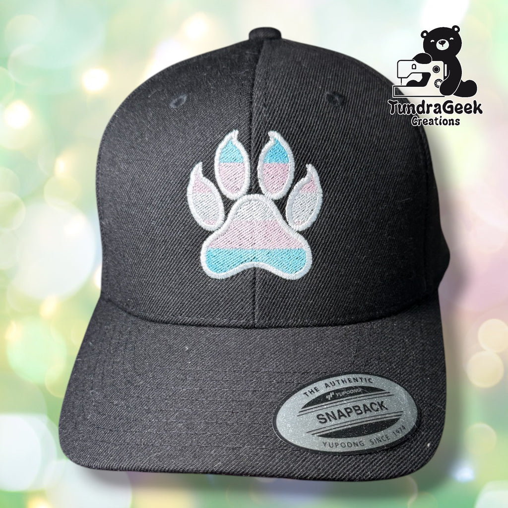 Black snapback cap with an embroidered trans pride paw print in white, pink, and blue. TundraGeek Creations logo in the top right corner.