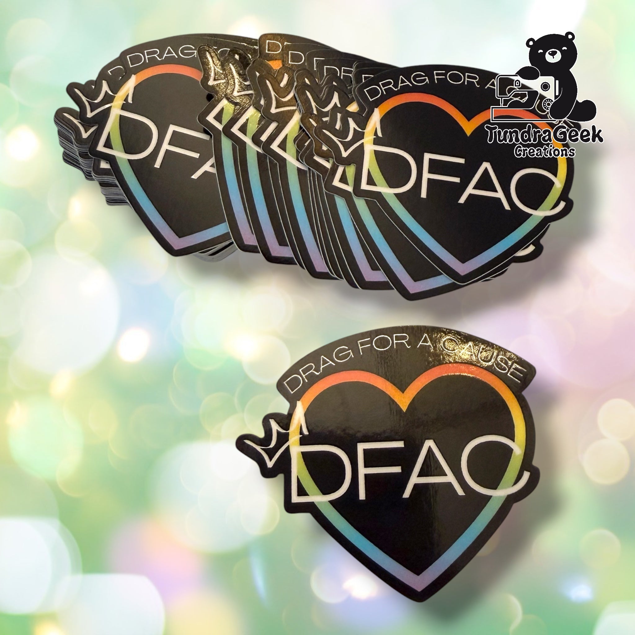 A set of black heart-shaped stickers with the text 'DRAG FOR A CAUSE' and 'DFAC' in white, outlined with a rainbow gradient border, shown on a soft green and gold bokeh background. A Tundra Geek Creations logo is visible in the top right corner.