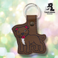 Front of a brown faux leather pibble keychain. An embroidered pit bull terrier stands facing left with a white chest blaze, red collar, red ear accents, and black nose and facial detail stitching on a matching brown vinyl base. A bronze 9mm metal rivet closure is visible on the tab, with a silver split ring above.
