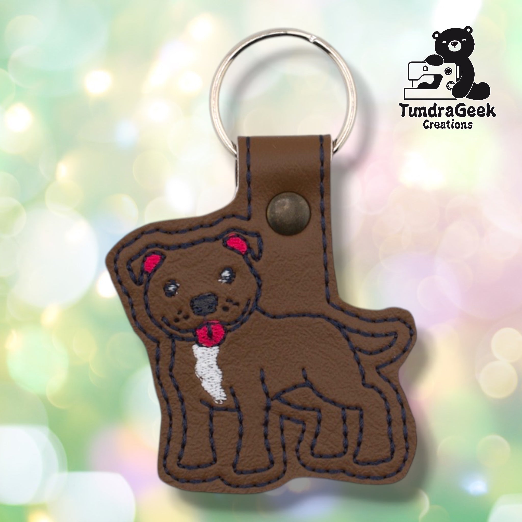Front of a brown faux leather pibble keychain. An embroidered pit bull terrier stands facing left with a white chest blaze, red collar, red ear accents, and black nose and facial detail stitching on a matching brown vinyl base. A bronze 9mm metal rivet closure is visible on the tab, with a silver split ring above.