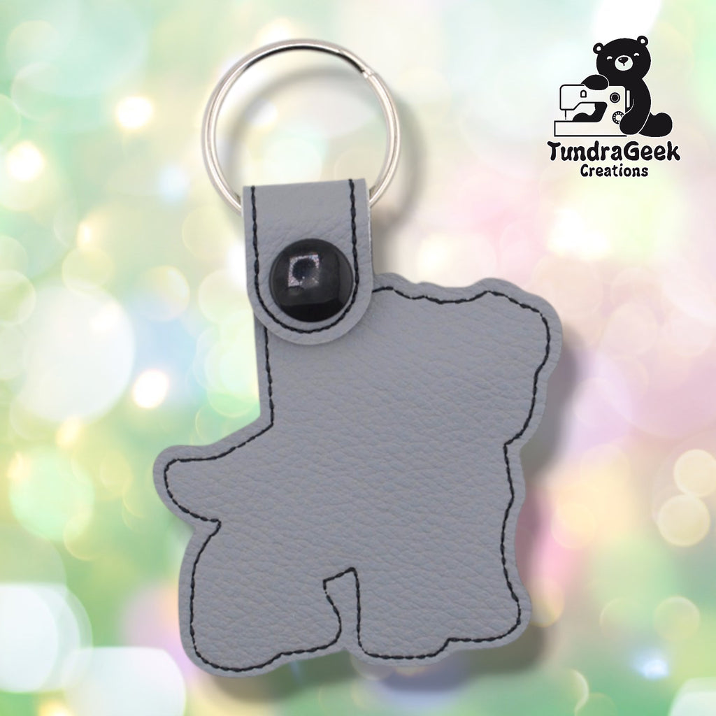 Back of a gray faux leather pibble keychain. The plain gray vinyl back panel shows dark outline stitching following the dog silhouette shape. A black 12.5mm plastic snap closure secures the tab, with a silver split ring attached above.