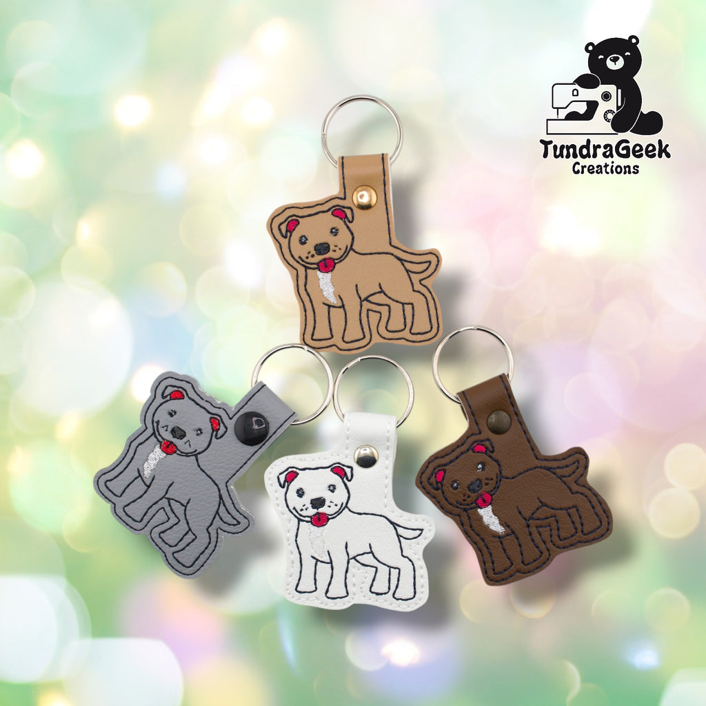 All four pibble keychain variants arranged together against a soft bokeh background. Tan is shown at top, gray at left, white at bottom center, and brown at right. Each features the same embroidered pit bull terrier design on a matching faux leather base.