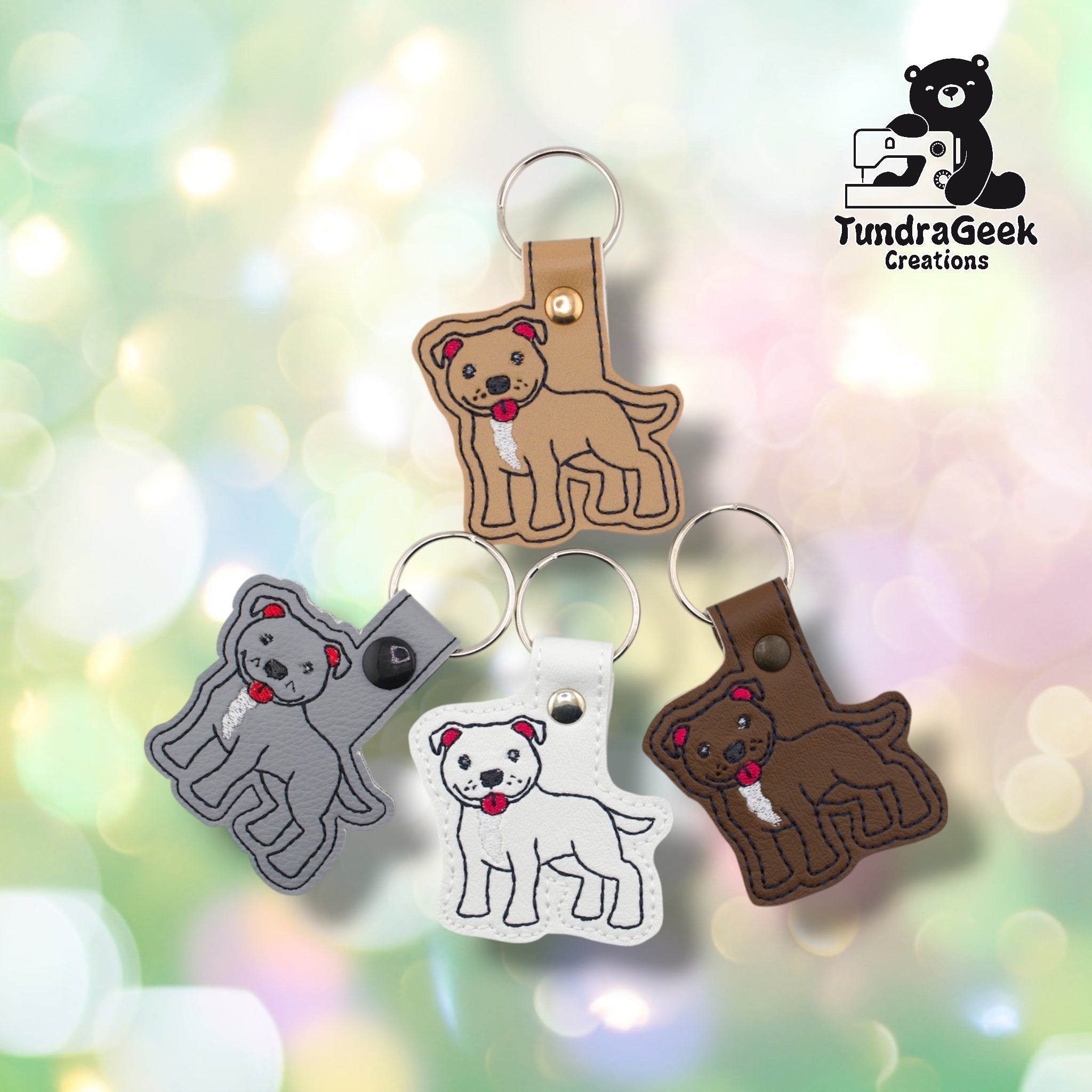 All four pibble keychain variants arranged together against a soft bokeh background. Tan is shown at top, gray at left, white at bottom center, and brown at right. Each features the same embroidered pit bull terrier design on a matching faux leather base.