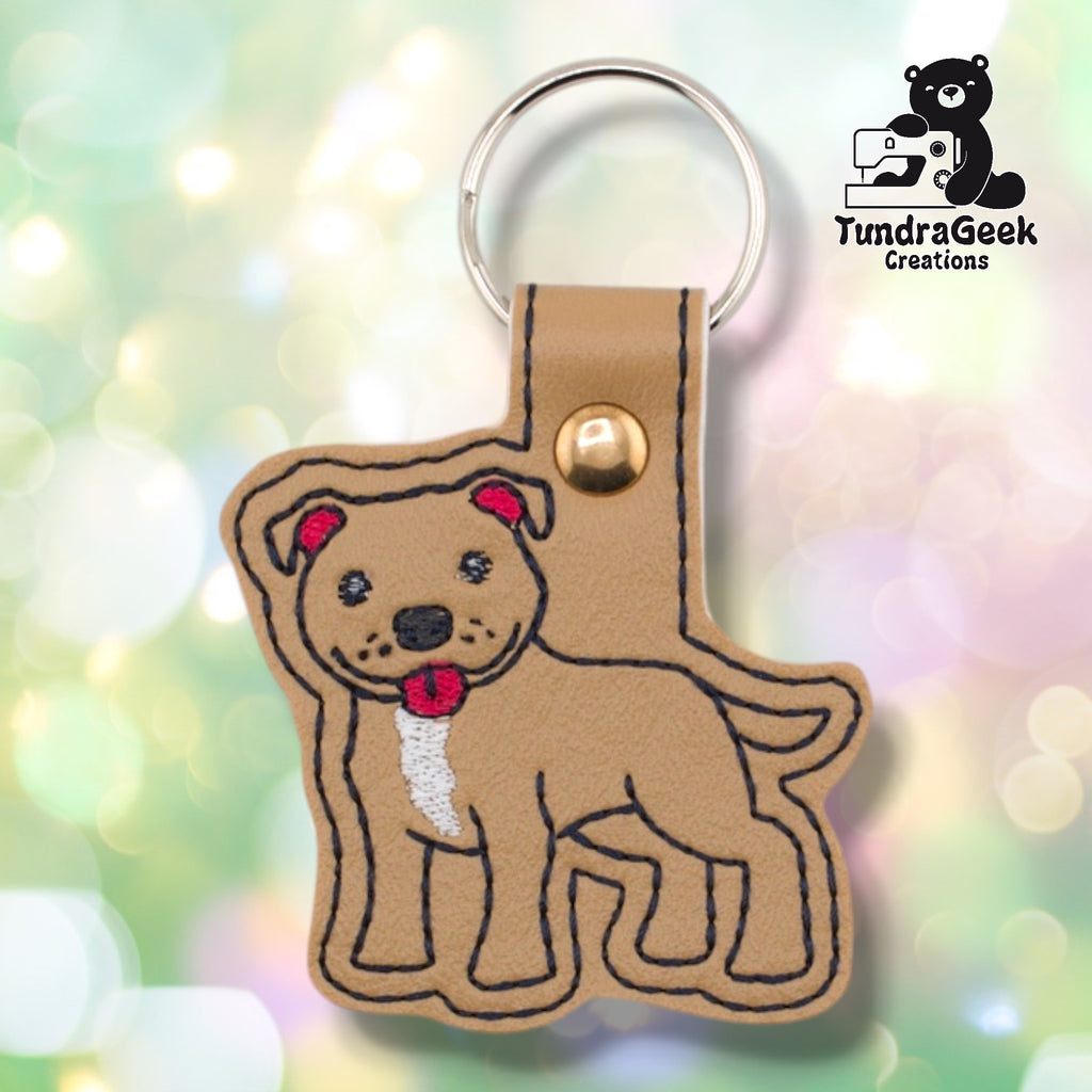 Front of a tan faux leather pibble keychain. An embroidered pit bull terrier stands facing left with a white chest blaze, red collar, red ear accents, and black nose and facial detail stitching on a matching tan vinyl base. A rose gold 9mm metal rivet closure is visible on the tab, with a silver split ring above.