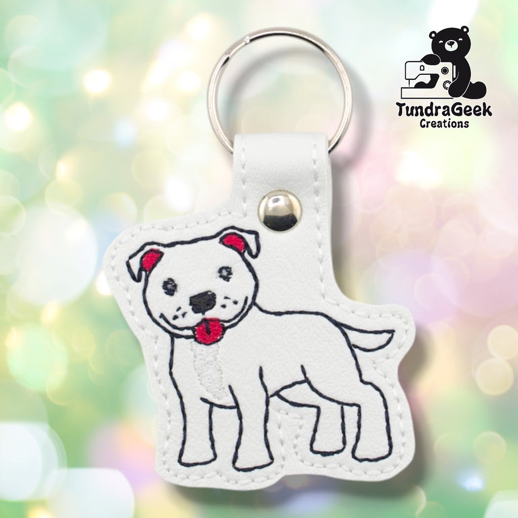 Front of a white faux leather pibble keychain. An embroidered pit bull terrier stands facing left with a white chest blaze, red collar, red ear accents, and black nose and facial detail stitching on a white vinyl base. A silver 9mm metal rivet closure is visible on the tab, with a silver split ring above.