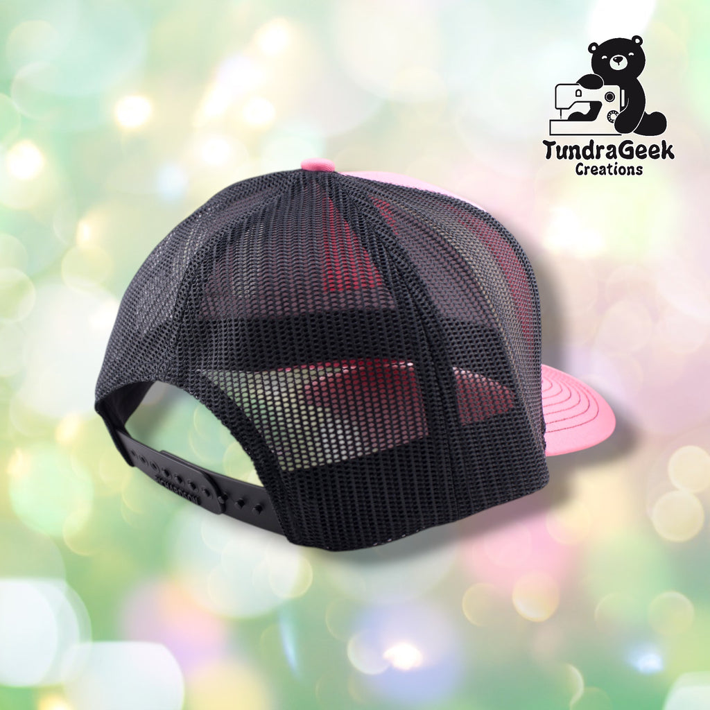 A black mesh trucker cap with pink bill displayed against a soft, blurred pastel background.
