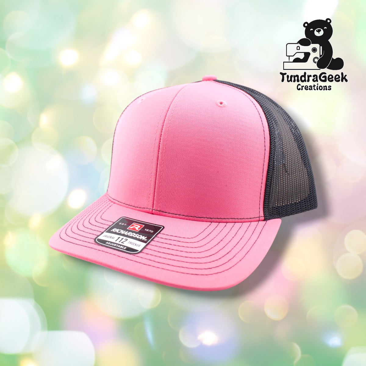 A pink and black mesh trucker cap with stitched details and an adjustable back label displayed against a soft bokeh background.