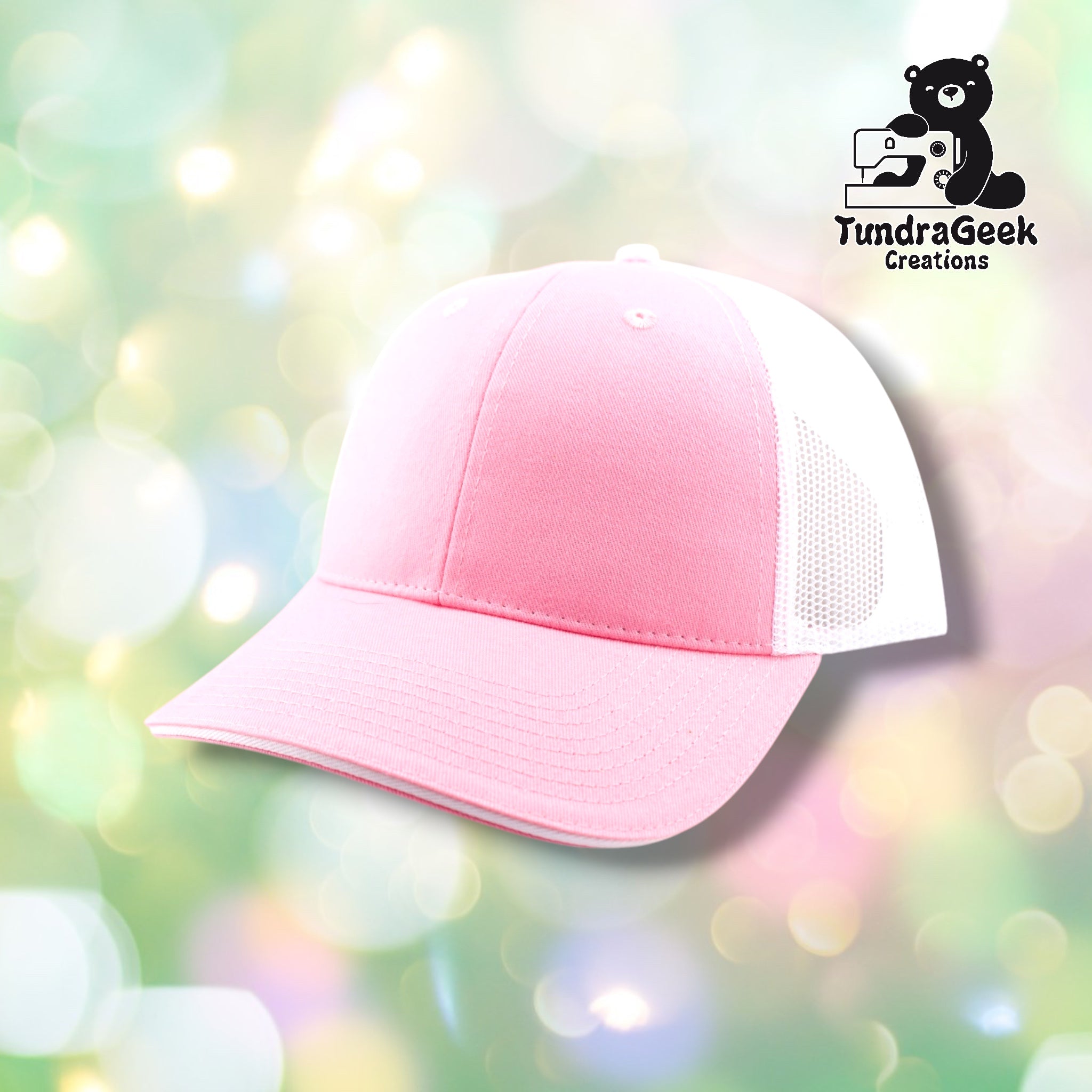A pink and white mesh trucker cap displayed against a soft, blurred pastel bokeh background with the TundraGeek Creations logo visible in the upper right.