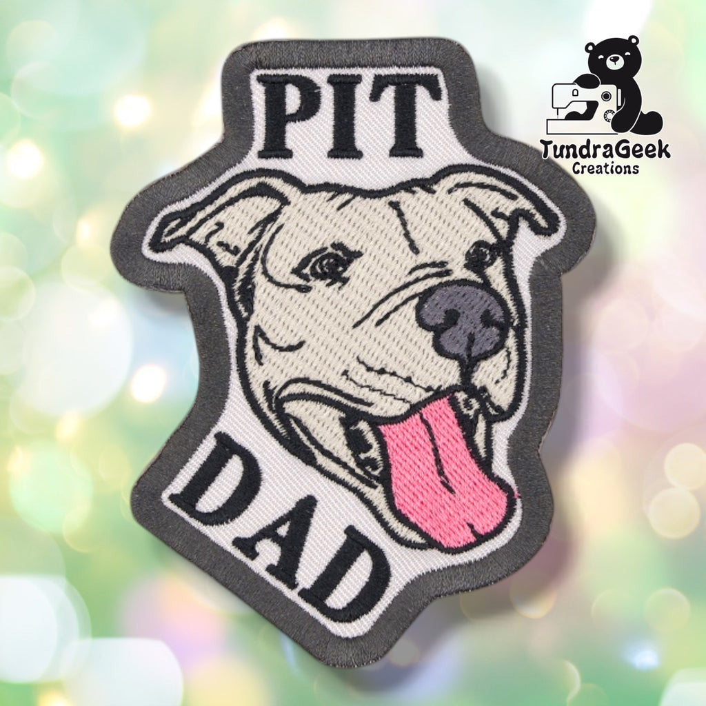 Grey embroidered patch with a black merrowed edge, showing a smiling full-eared pitbull with its tongue out. "PIT" is stitched above the dog's head and "DAD" below, both in black block lettering.