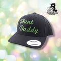 A black snapback cap with green embroidered script reading "plant Daddy" on a soft-focus green and white bokeh background.