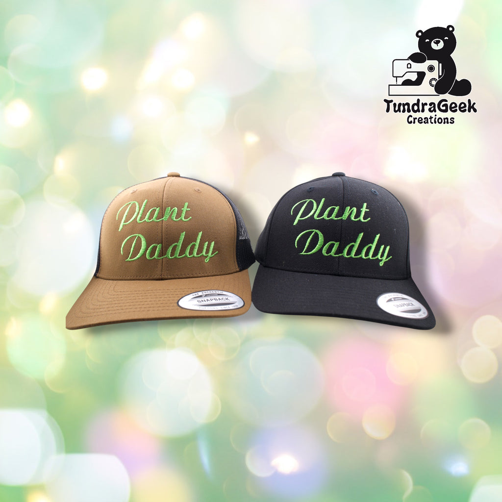 Two trucker hats with mesh backs displayed against a blurred green and white bokeh background; one tan and one black, both embroidered with "Plant Daddy" in green script.
