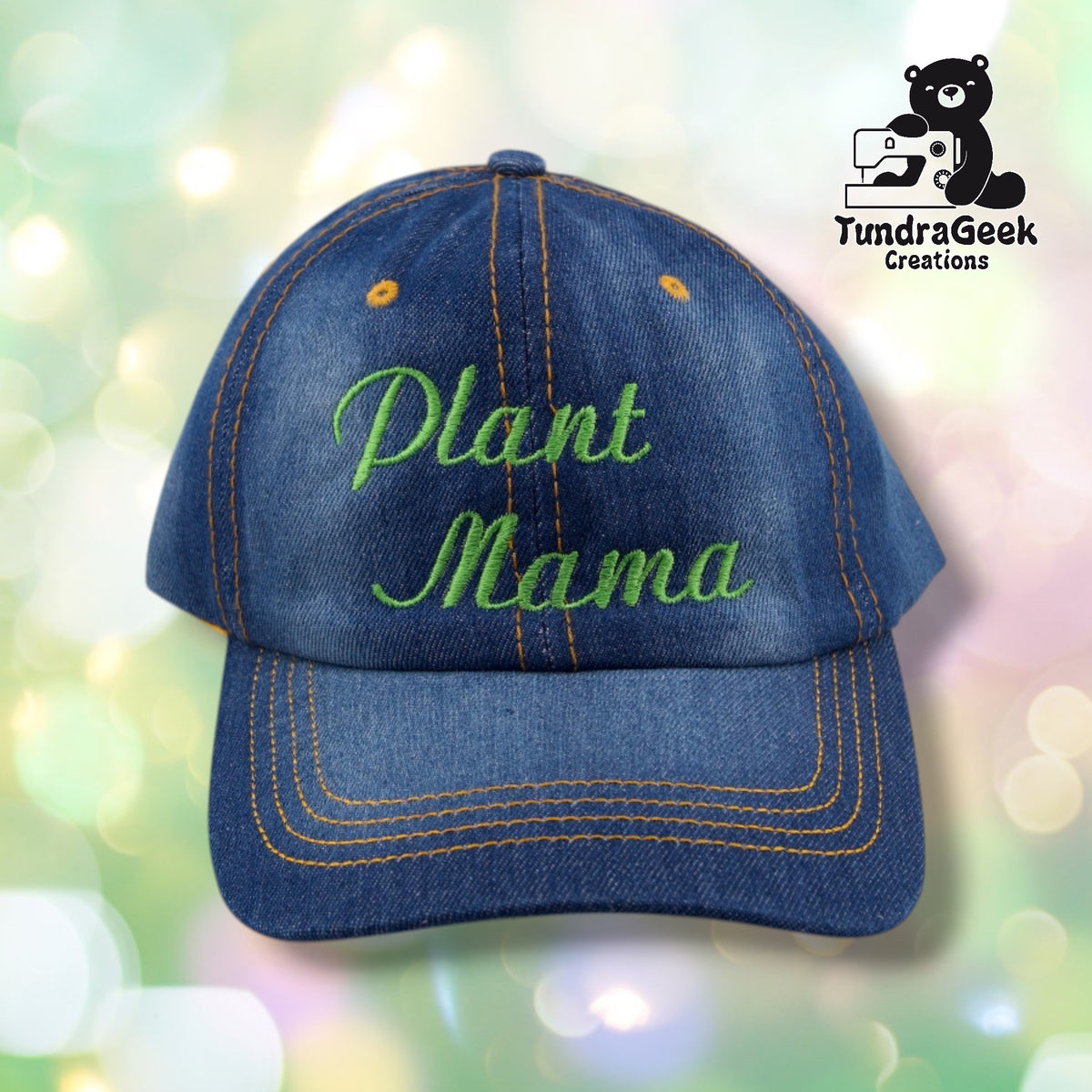 A denim baseball cap with orange stitching and green embroidered "Plant Mama" text on a blurred green and white bokeh background.