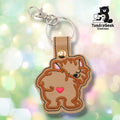 Tan faux leather keychain shaped like a Pomeranian viewed from behind. Brown embroidery details the fluffy fur around the body; small pink inner ears are visible over the shoulders and a sleepy eye peeks around one side. A small pink heart is embroidered on the center of the dog's rear. Gold metal rivet closure and silver key ring at top. TundraGeek Creations logo in upper right corner.