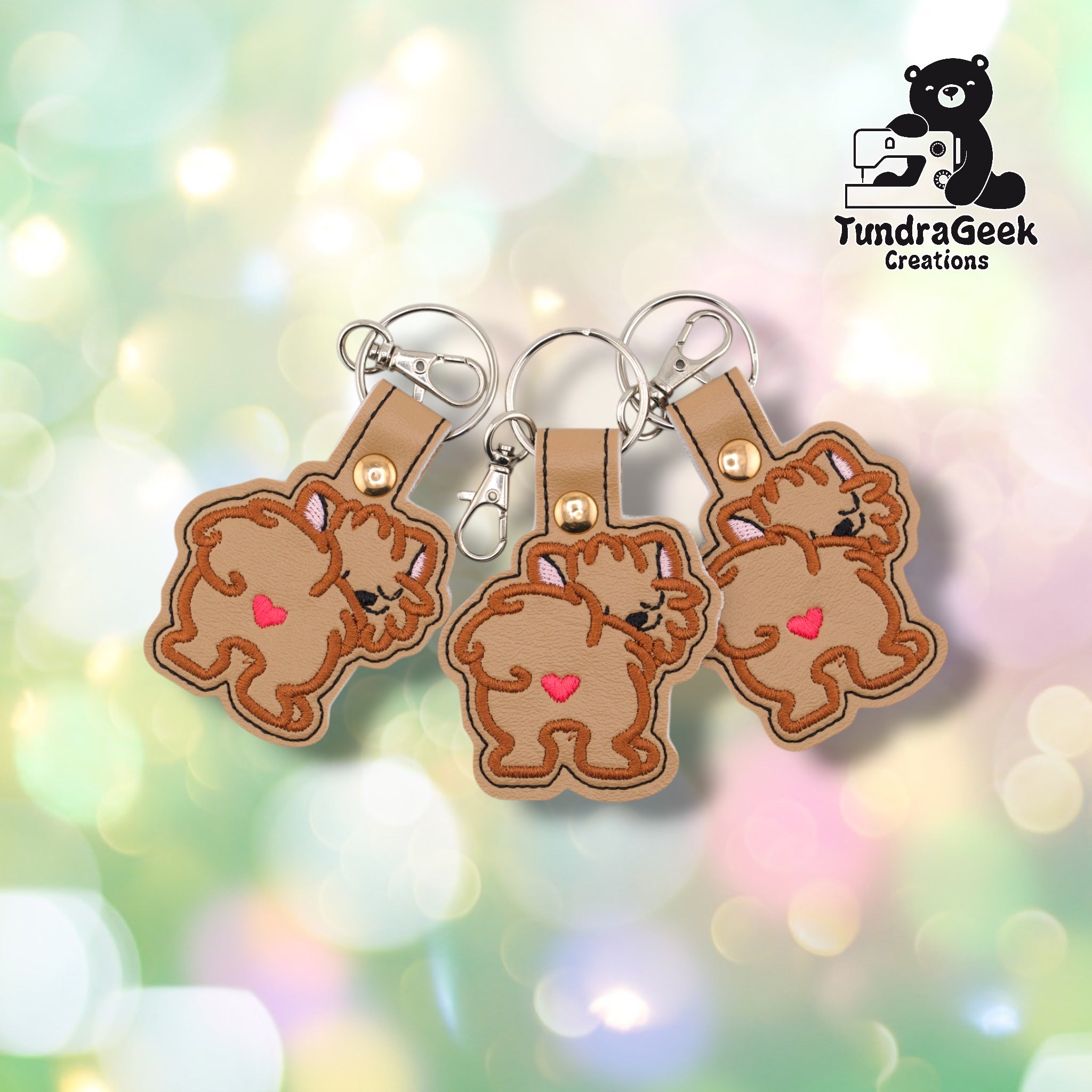 Three Pomeranian keychains fanned out on a pastel bokeh background, showing the fluffy rear design from slightly varied angles. All three feature matching brown embroidery, tan base, and gold metal rivet closures.