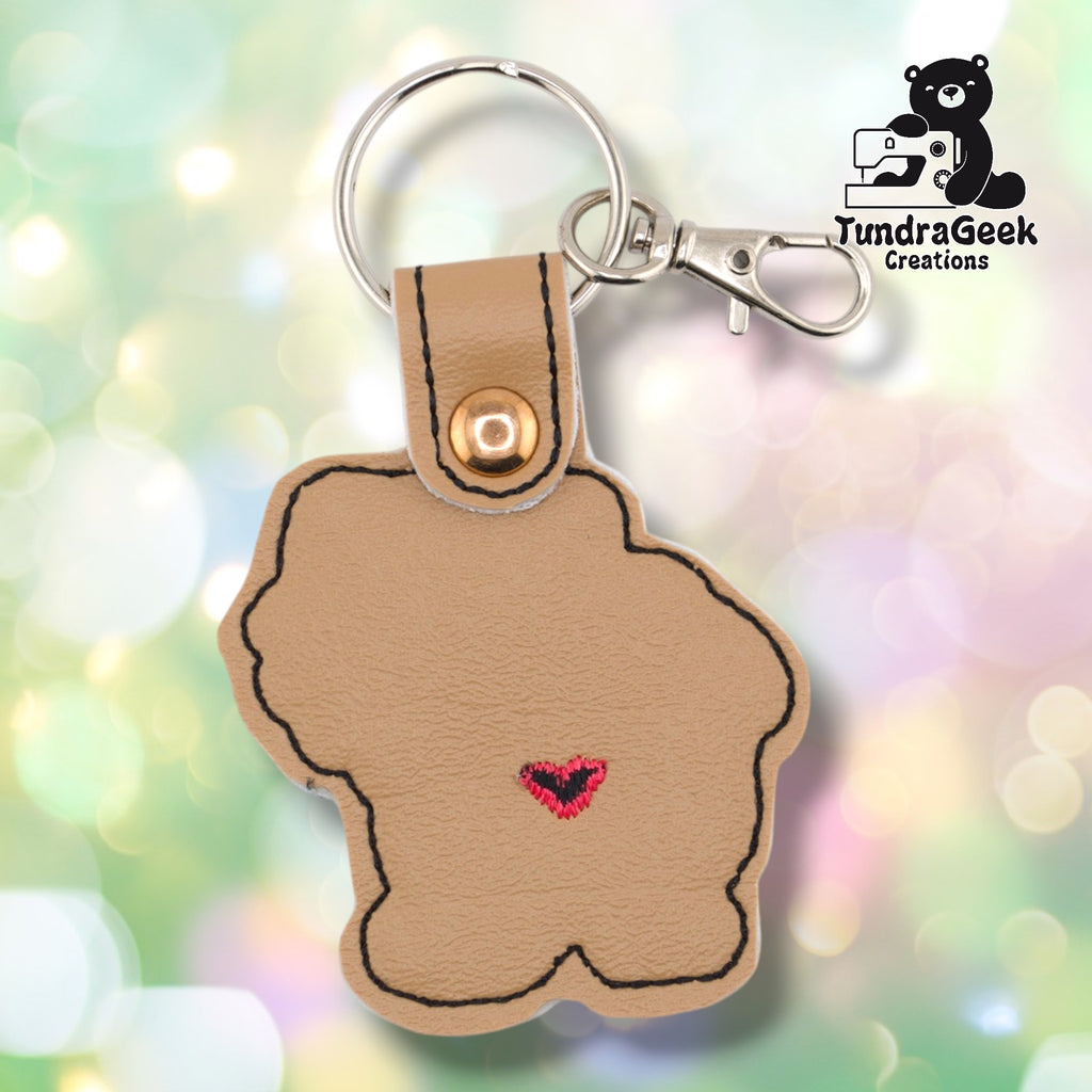 Plain back of the Pomeranian keychain showing tan faux leather and black outline stitching following the rounded fluffy silhouette. A small pink heart is also embroidered on this side. Gold metal rivet closure visible at top.