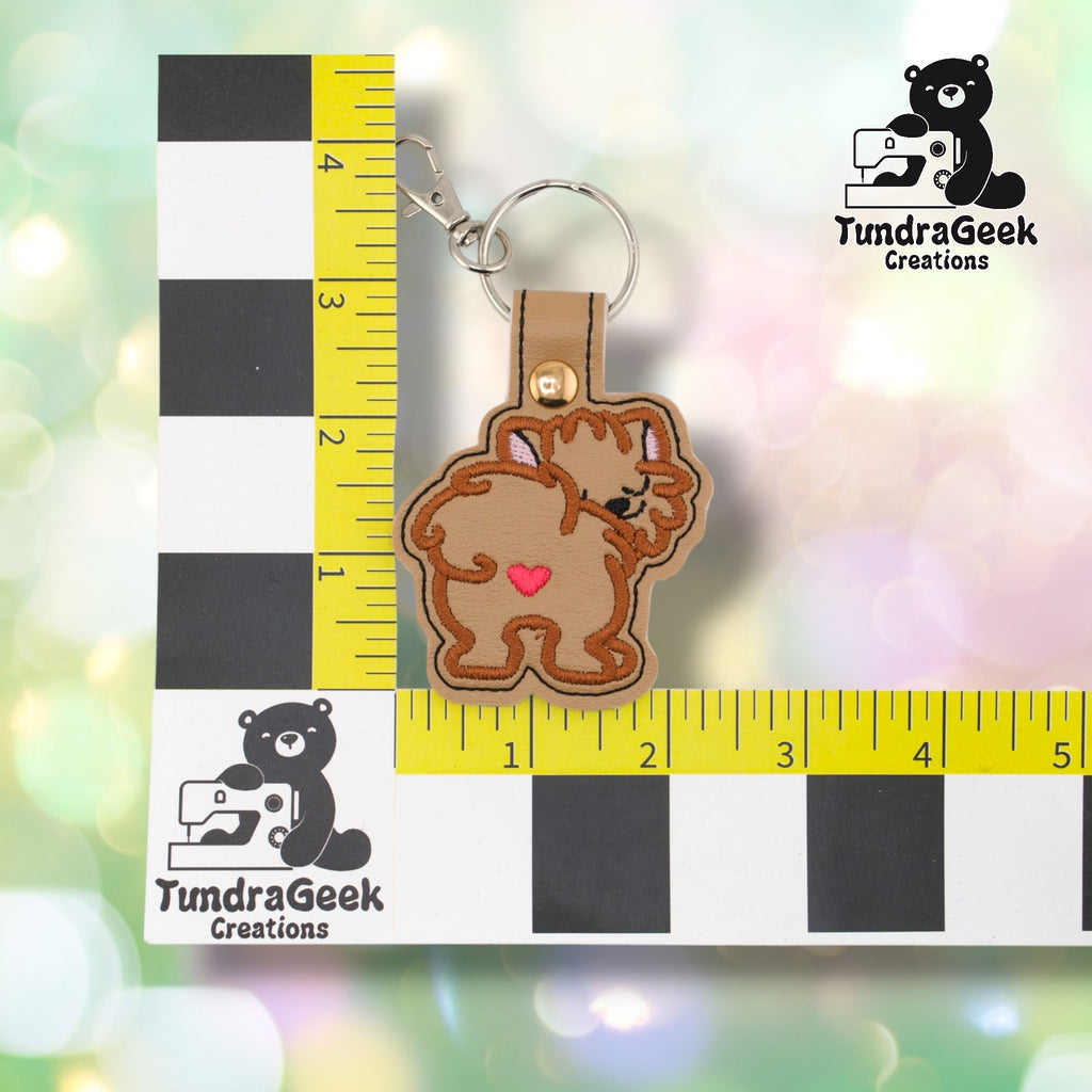 Pomeranian keychain positioned against a TundraGeek Creations branded ruler card showing yellow tape measure. Keychain body measures approximately 2.5 inches wide by 2.25 inches tall, not including hardware.