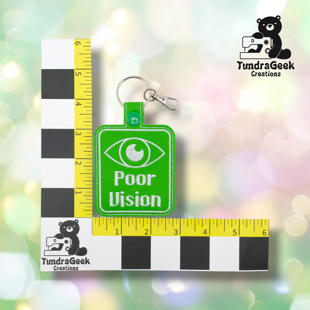 A green keychain with an eye graphic and the text Poor Vision is placed on a black and white ruler grid for scale, with a TundraGeek Creations logo in the corners.