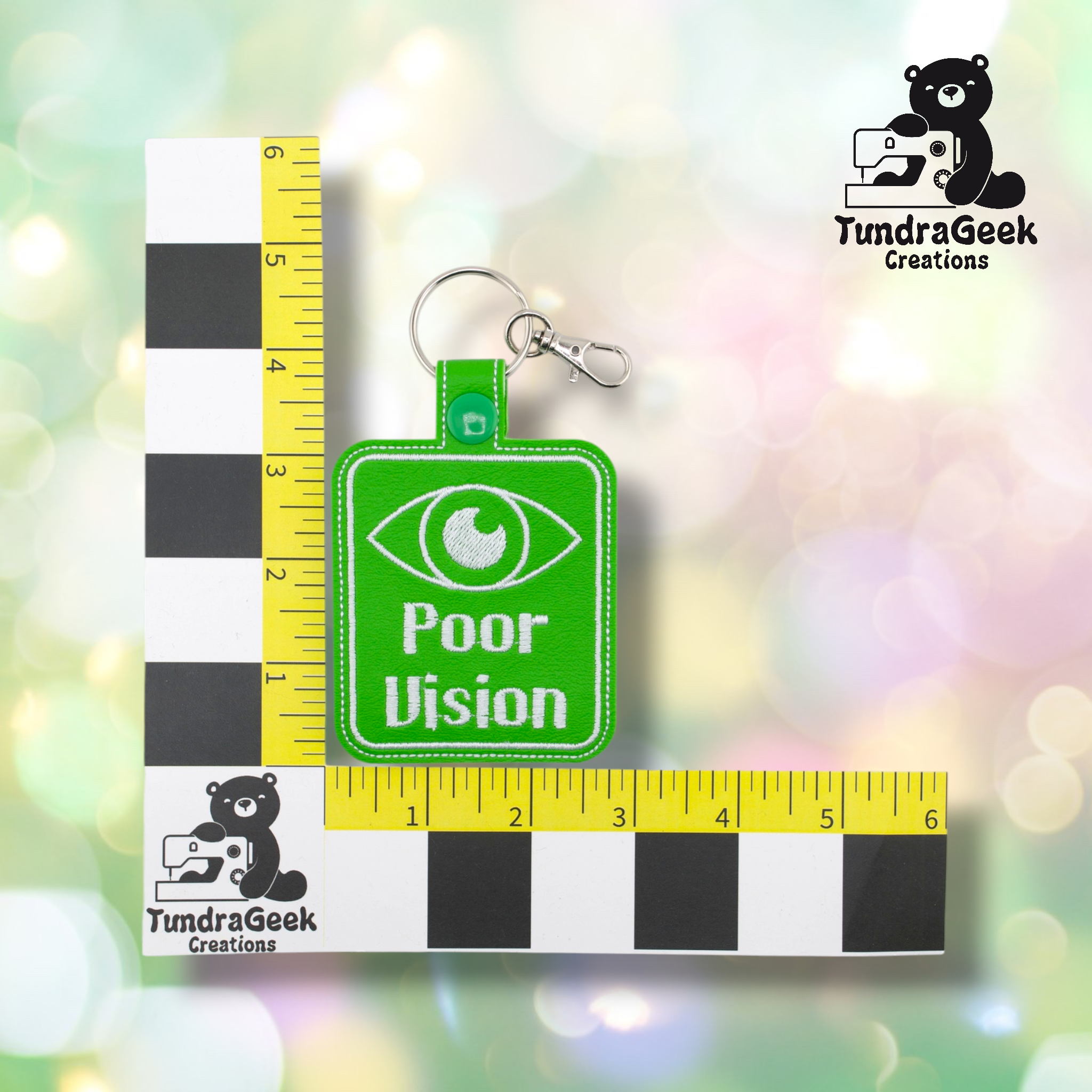 A green keychain with an eye graphic and the text Poor Vision is placed on a black and white ruler grid for scale, with a TundraGeek Creations logo in the corners.