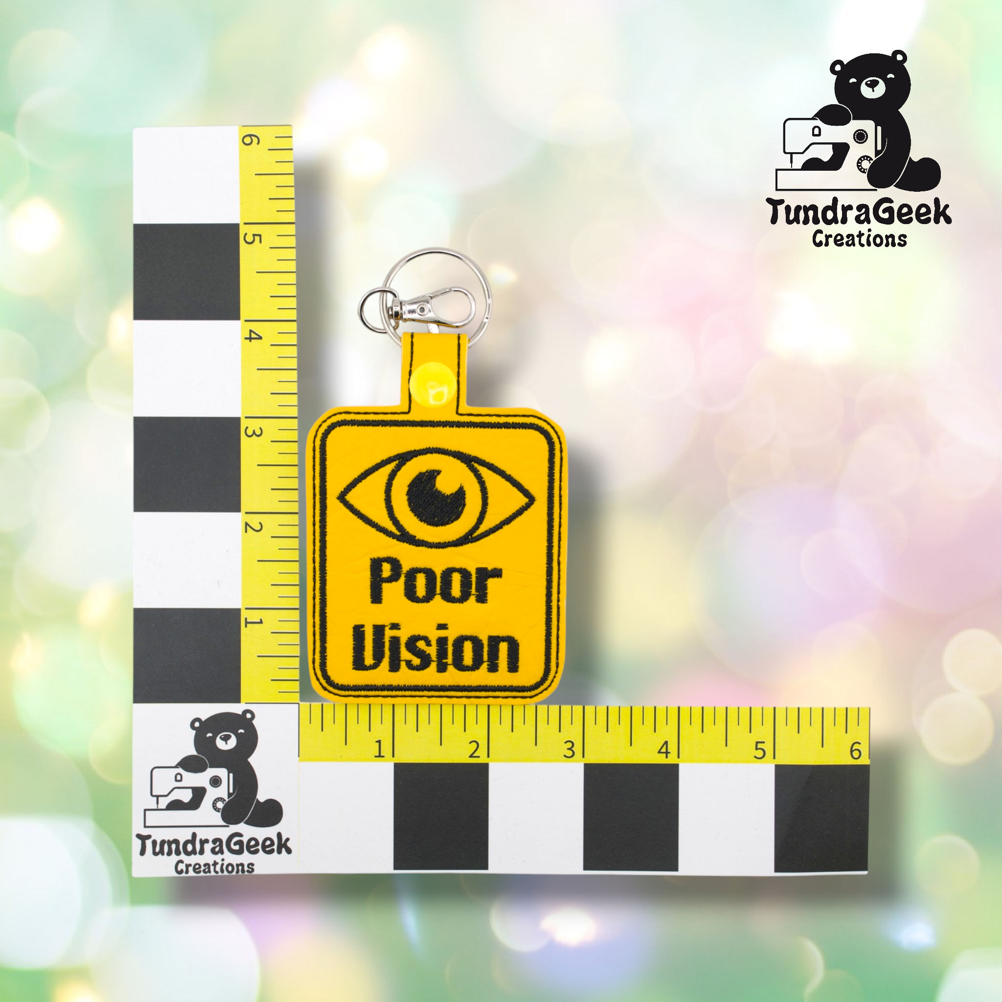 Yellow keychain with a black eye graphic and the words Poor Vision is placed on a measurement grid with TundraGeek Creations logos in the corners.