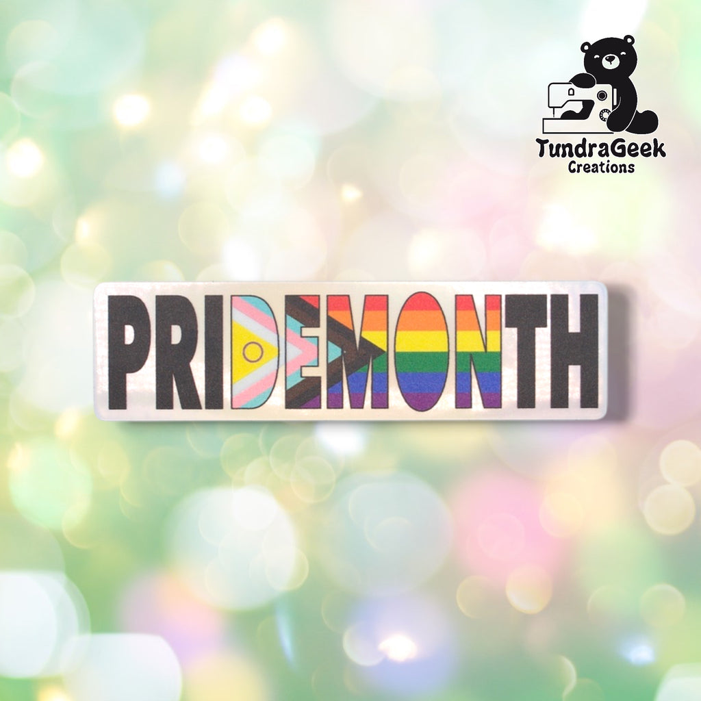 Horizontal die-cut holographic sticker reading "PRIDEMONTH" in bold block lettering on a silver holographic background. The letters D, E, M, O, and N are filled with Intersex Progress Pride flag colors; remaining letters display classic rainbow stripes. TundraGeek Creations logo in upper right corner.