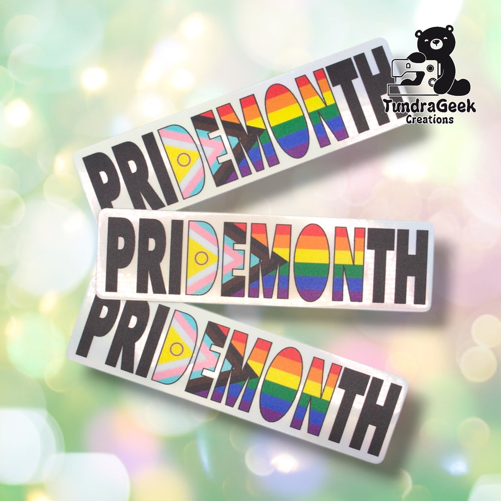 Three Pride Month holographic stickers fanned out on a pastel bokeh background, showing the design from slightly varied angles. All three feature the same holographic silver background and multicolor pride flag lettering.